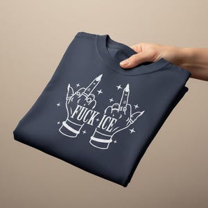May include: Dark navy blue t-shirt featuring a white graphic of two hands giving the middle finger, with the text "FUCK + ICE". The design includes stars and jewellery details. The shirt is folded.