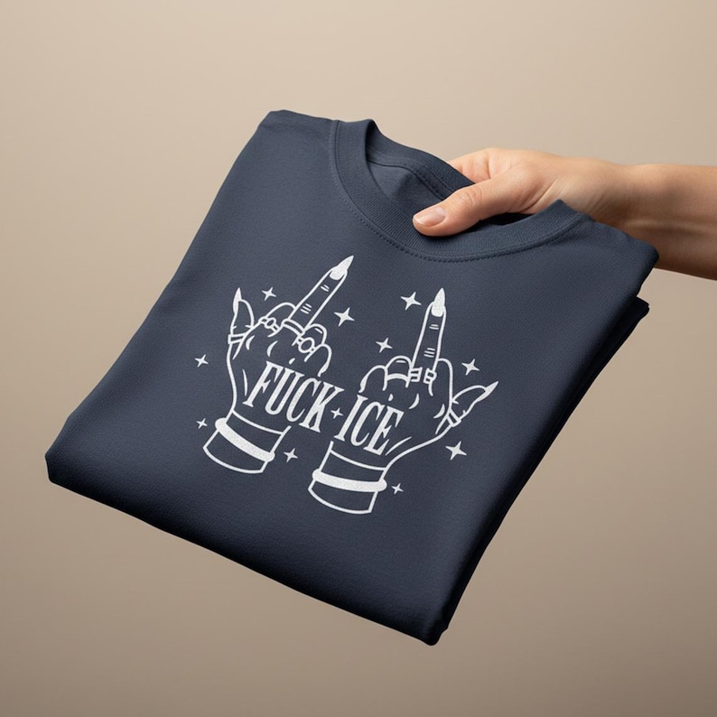 May include: Dark navy blue t-shirt featuring a white graphic of two hands giving the middle finger, with the text "FUCK + ICE". The design includes stars and jewellery details. The shirt is folded.