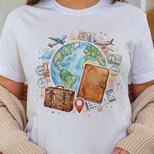 May include: White t-shirt featuring a watercolor design of a globe, airplanes, stamps, a suitcase, and a passport with the text "ADVENTURE AWAITS". The design includes a red location marker and travel-themed elements.