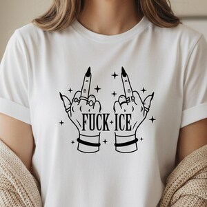 May include: White t-shirt featuring a black graphic of two hands displaying the middle finger, embellished with rings and bracelets. The text "FUCK ICE" is printed below, with star accents.