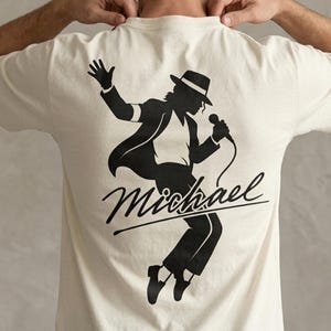 Puede incluir: Camiseta color crema con una silueta negra de un cantante con fedora y micr&oacute;fono. La palabra "Michael" est&aacute; escrita en cursiva debajo. El dise&ntilde;o est&aacute; centrado en la parte posterior.
