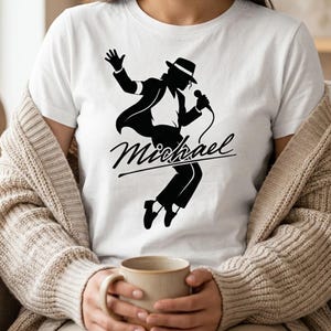 Puede incluir: Camiseta blanca con una silueta negra de un artista con sombrero y traje, sosteniendo un micr&oacute;fono. El nombre "Michael" est&aacute; escrito en cursiva debajo. La camiseta es de cuello redondo cl&aacute;sico.