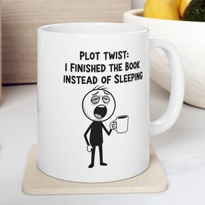 May include: White ceramic mug with the text "PLOT TWIST: I FINISHED THE BOOK INSTEAD OF SLEEPING" and a cartoon figure holding a mug. The figure has a tired expression.