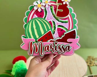 Può includere: Un cake topper a tema anguria con il nome "Marissa" e il numero "5". Il topper presenta un design di anguria verde e rossa, fiori bianchi e uno striscione rosa. Lo sfondo è verde unito.