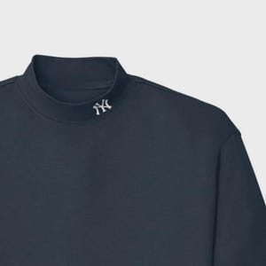 May include: A navy blue mock neck shirt with a white embroidered logo on the upper chest. The shirt has a simple, classic design and is made of a soft material.