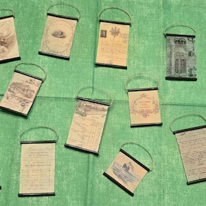 May include: A collection of vintage-style hanging scrolls with various designs, including illustrations, stamps, and handwritten text. Each scroll has a black frame and a metal handle for hanging. The scrolls are arranged on a green fabric surface.