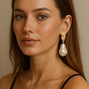 May include: Gold-coloured earrings featuring a textured top section and a large, pear-shaped pearl drop. The earrings are worn by a person with brown hair and a black top. The background is a neutral beige.