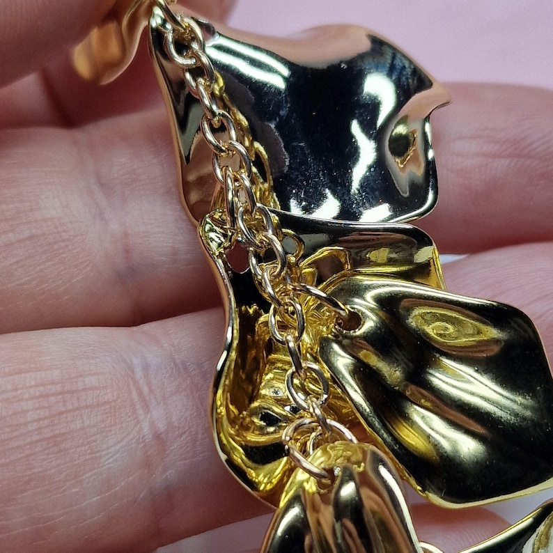 May include: Gold-coloured statement earrings with a layered, petal-like design. The earrings have a high-shine finish and are connected by a delicate chain. The design is abstract and modern, suitable for special occasions.
