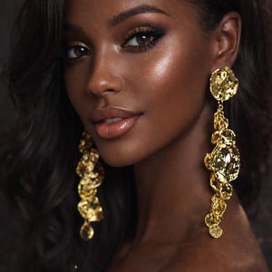 May include: A portrait of a person with dark skin, wearing long, dangling gold earrings. The earrings have an intricate, textured design. The person has dark hair and is wearing makeup, including eyeshadow and lip gloss.