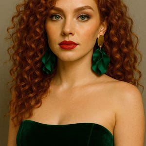 Op de afbeelding: Een vrouw met rood krullend haar en groene ogen draagt een donkergroene strapless fluwelen top en grote groene, bladvormige oorbellen met gouden schijfjes. Haar lippen zijn rood geschilderd.