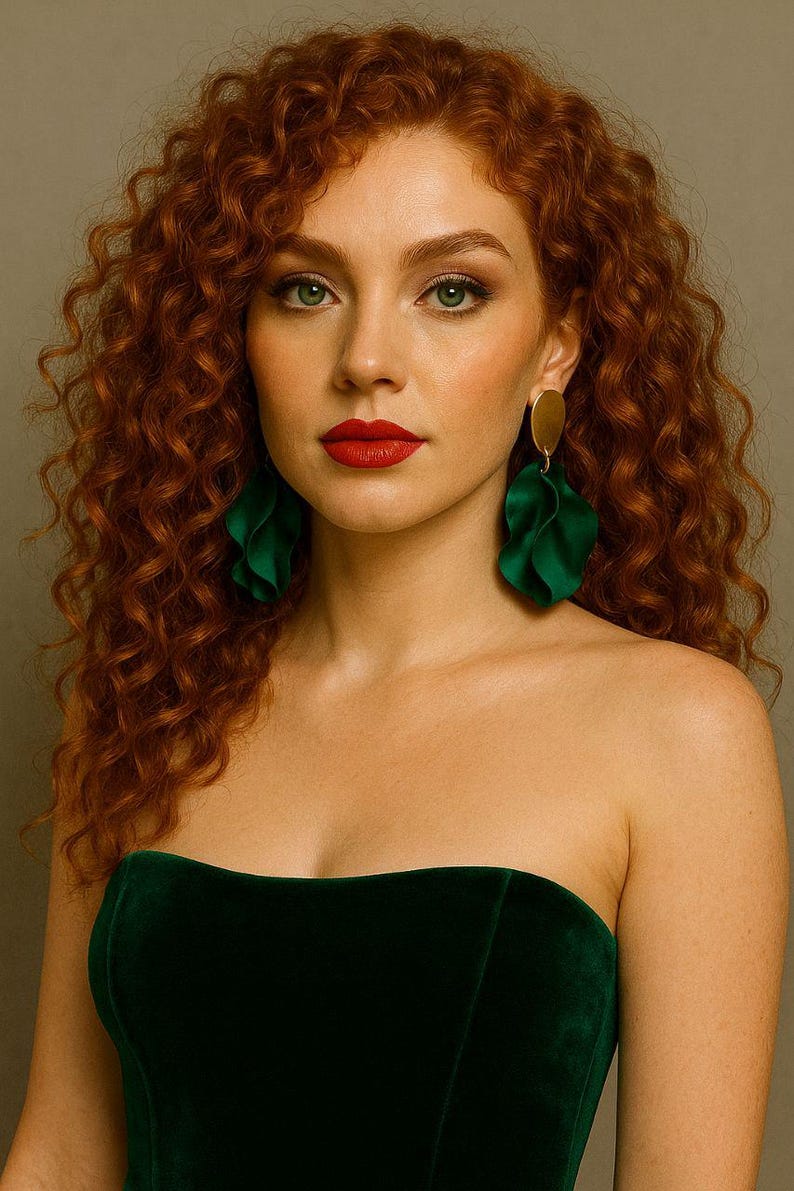 Op de afbeelding: Een vrouw met rood krullend haar en groene ogen draagt een donkergroene strapless fluwelen top en grote groene, bladvormige oorbellen met gouden schijfjes. Haar lippen zijn rood geschilderd.