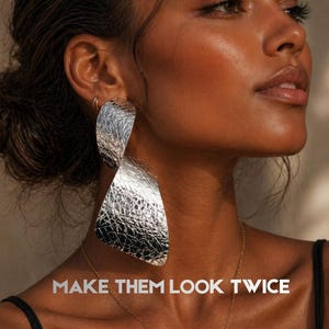 Puede incluir: Una mujer de piel oscura lleva pendientes grandes y plateados con textura. Los pendientes tienen una forma abstracta. Se ve un collar de oro. El texto "MAKE THEM LOOK TWICE" se muestra en letras plateadas.