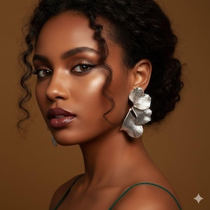 Op de afbeelding: Een vrouw met donker krullend haar en donkere lippenstift draagt grote, zilveren, blad-vormige oorbellen. De oorbellen hebben een getextureerde, metallic afwerking en hangen aan haar oren. Ze draagt een groene top met spaghettibandjes.