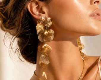 Puede incluir: Pendientes dorados con diseño floral en cascada. Los pendientes presentan múltiples capas de delicados pétalos texturizados que cuelgan con gracia. Un collar dorado a juego con un colgante floral complementa los pendientes, creando un conjunto de joyería.