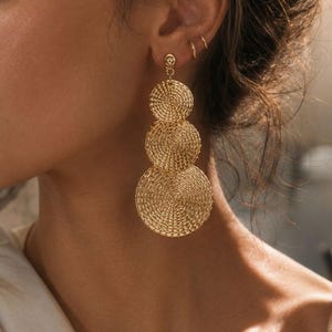 May include: A gold-toned drop earring featuring three circular discs of varying sizes arranged vertically. Each disc has a detailed, concentric, wire-like textured pattern. The earring is attached to the earlobe with a small, simple gold stud component.