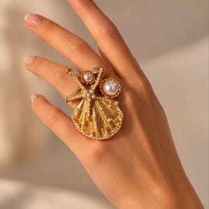 May include: A gold ring with a seashell design, adorned with pearls of varying sizes. The ring is displayed on a hand, highlighting its intricate details and elegant appearance. The ring's design incorporates a fan-shaped element.