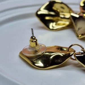 May include: Close-up of gold-coloured earrings with a leaf-like design and a shiny, reflective surface. The earring post and backing are visible, showing the details of the jewelry's construction. The earrings are on a white surface.