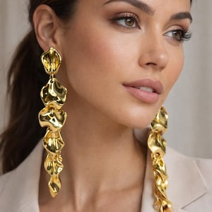 Puede incluir: Pendientes largos colgantes dorados. Cada pendiente presenta una serie de elementos texturizados en forma de pétalo que caen en cascada. Los pendientes son de un oro brillante y parecen ser de metal. La persona lleva una blazer color crema.