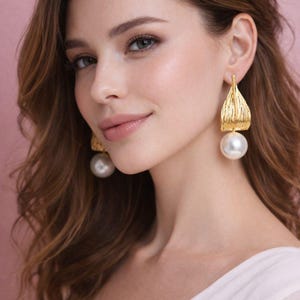 Puede incluir: Pendientes dorados con una parte superior texturizada en forma de hoja y una gran gota de perla blanca redonda. Los pendientes los lleva una persona con cabello castaño y una blusa blanca, sobre un fondo rosa.