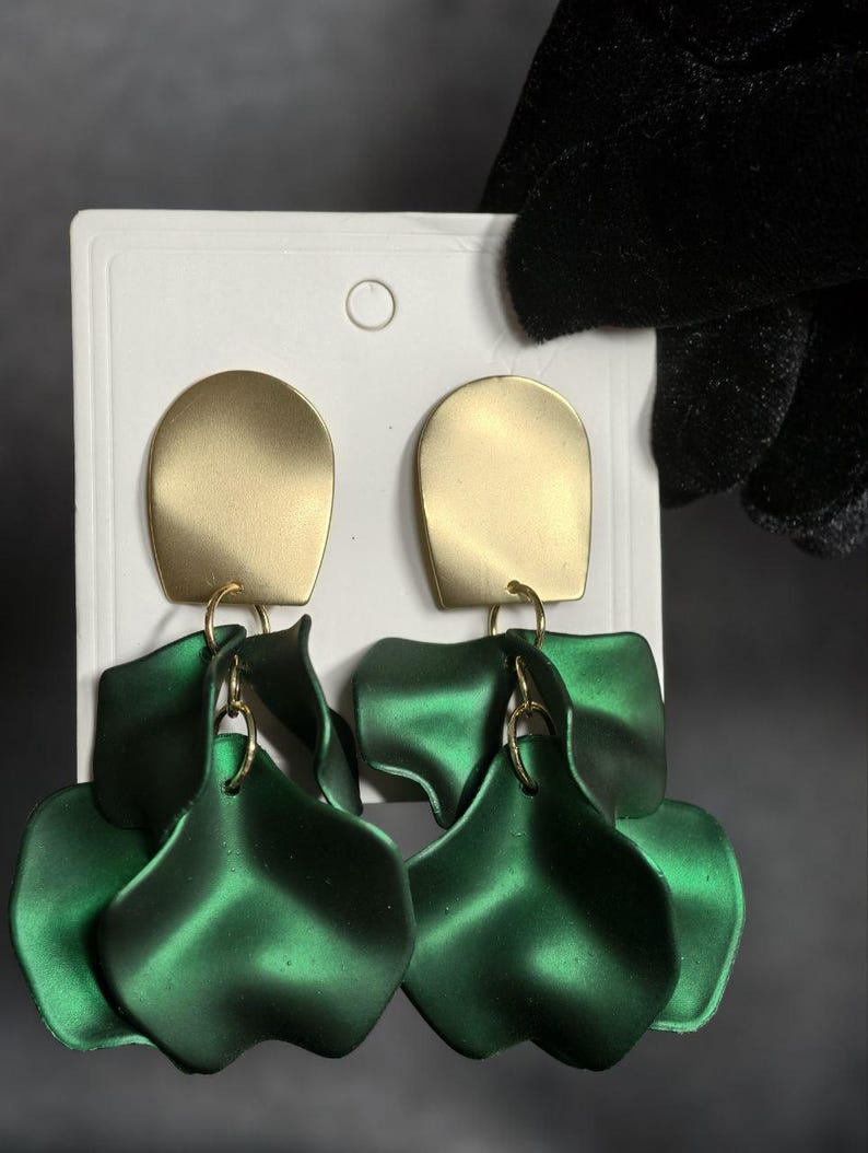Op de afbeelding: Een paar goudkleurige, rechthoekige oorbellen met een matte afwerking. Daaronder hangen drie gelaagde, bloemblad-vormige, smaragdgroene oorbellen aan goudkleurige ringen. De oorbellen worden gepresenteerd op een witte kaart.
