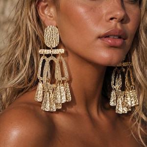 May include: Gold-coloured statement earrings with a textured, hammered metal finish. The earrings feature an oval top, a horizontal bar, arched shapes, and dangling, bell-shaped elements. These bold earrings are a striking accessory.