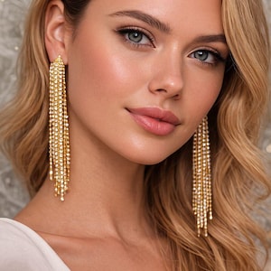 Könnte beinhalten: Nahaufnahme einer Person, die goldfarbene, baumelnde Ohrringe trägt. Die Ohrringe bestehen aus mehreren Strängen kleiner, funkelnder Kristalle. Die Person hat blondes Haar und trägt ein weißes Oberteil. Die Ohrringe sind ein glamouröses Accessoire.