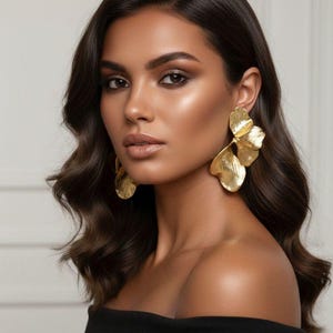 Puede incluir: Una mujer con cabello oscuro y ondulado y pendientes dorados en forma de hoja. Lleva un top negro con los hombros descubiertos. Los pendientes son grandes y texturizados, captando la luz. Su piel tiene un tono cálido y brillante.