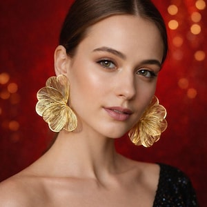 May include: A woman wearing large, gold-coloured statement earrings. The earrings have a layered, petal-like design. The background is a blurred red with bokeh lights.
