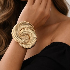 May include: A gold-colored cuff bracelet with a spiral design. The bracelet is made of a shiny metal and has a textured surface. The jewelry is worn on the wrist and is a statement piece.
