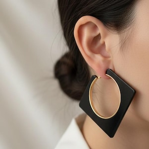 May include: Black square earrings with a gold hoop design. The earrings are worn on an ear, with the square portion framing the earlobe. The earrings have a modern, geometric design.
