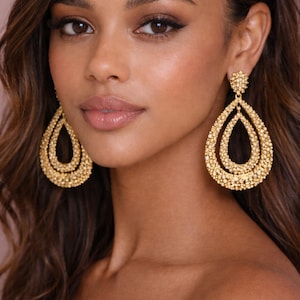 May include: Close-up of a person wearing large, gold-coloured teardrop earrings. The earrings have a textured, beaded appearance and a layered design. The earrings are a statement piece, suitable for special occasions.