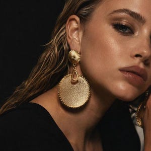 May include: A pair of gold-toned drop earrings featuring a textured circular pendant suspended from a smaller, ribbed, dome-shaped stud. The large circular disc has a granulated surface finish and a beaded edge, connected by a small teardrop link.