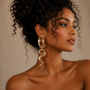 May include: A woman with dark curly hair is wearing gold-coloured, abstract drop earrings. The earrings have a unique, flowing design with a loop at the top and a curved, elongated shape. The woman's skin has a warm tone, and she is wearing a cream-coloured top.