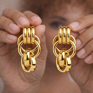 May include: A pair of gold-colored earrings. Each earring features a series of connected, rounded shapes, including three stacked bars at the top, a large ring, and two smaller, rounded shapes hanging below. The earrings are held up by a person's hands.