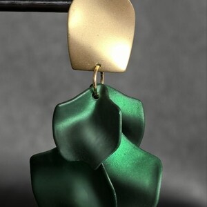 Op de afbeelding: Een goudkleurige, gebogen rechthoekige oorbel met drie gelaagde, donkergroene, bloemblad-vormige hangers. De oorbel hangt aan een zwarte staaf tegen een grijze achtergrond. De sieraden hebben een modern, minimalistisch ontwerp.