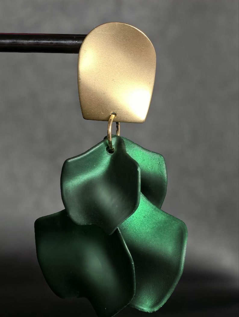 Op de afbeelding: Een goudkleurige, gebogen rechthoekige oorbel met drie gelaagde, donkergroene, bloemblad-vormige hangers. De oorbel hangt aan een zwarte staaf tegen een grijze achtergrond. De sieraden hebben een modern, minimalistisch ontwerp.