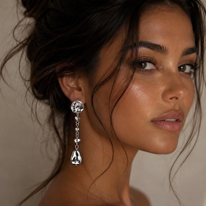 May include: A close-up of a woman wearing a silver earring. The earring features a floral design at the top, with a chain of small beads and a teardrop-shaped pendant. The woman has dark hair and is wearing a black top.