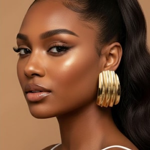 May include: A person with dark hair in a ponytail wears large, gold, rectangular earrings with a ridged design. The person has dark skin and is wearing a white top. The background is a neutral tan colour.