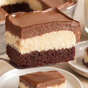 May include: A slice of layered chocolate dessert with a dark brown brownie base, a creamy vanilla middle layer, and a smooth chocolate mousse topping. A serving is lifted on a spatula, with another slice on a white plate.