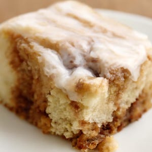 May include: A close-up of a cinnamon roll on a white plate. The pastry has a light brown swirl and is topped with a thick, white glaze. The roll is a square shape and appears soft and fluffy.