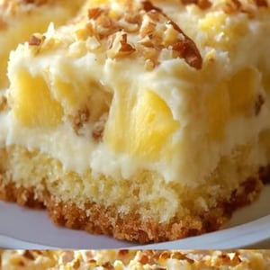 Peut inclure: Gros plan sur une part de gâteau à l'ananas. Le gâteau a une croûte brun doré, une couche de morceaux d'ananas jaunes et une garniture blanche crémeuse parsemée de noix hachées. Le gâteau est sur une assiette blanche.