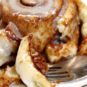 May include: A close-up of a cinnamon roll with a swirl design, topped with a white glaze. The roll is on a white plate with a fork. The pastry has a golden-brown color with visible layers and a glossy finish.