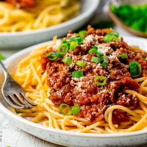 May include: A plate of spaghetti topped with a rich, red meat sauce, garnished with chopped green onions and grated cheese. The pasta is served in a white bowl with a silver fork. A second bowl of pasta is visible in the background.