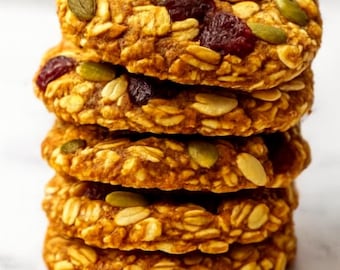 May include: A stack of five round, golden-brown cookies. Each cookie is studded with visible oats, pumpkin seeds, and dried cranberries. The cookies are stacked on top of each other, creating a visually appealing arrangement.