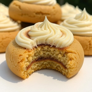May include: Close-up of several golden-brown cookies topped with swirls of white icing. One cookie has a bite taken out, revealing a dark filling. The cookies are arranged on a white surface, with a blurred green background.