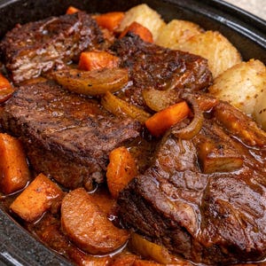 Könnte beinhalten: Nahaufnahme eines langsam gekochten Gerichts in einer schwarzen ovalen Schüssel. Das Gericht enthält große Stücke geschmortes Fleisch, umgeben von Stücken aus orangenen Karotten, Kartoffeln und Zwiebeln, alles in einer reichhaltigen, dunklen Soße.