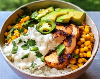 May include: A colorful bowl filled with grilled chicken, avocado slices, corn, and a dollop of white sauce. Garnished with cilantro and a jalapeño slice. The bowl sits on a light-colored surface.