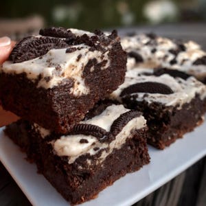 May include: Close-up of several chocolate brownies with a white cream topping and pieces of chocolate sandwich cookies. The brownies are stacked on a white plate, with a blurred background.