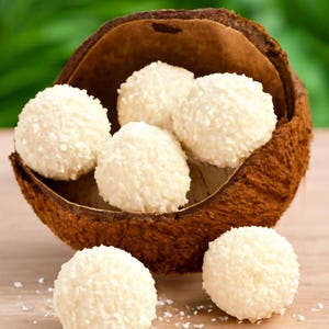 Puede incluir: Primer plano de caramelos redondos y blancos de coco, recubiertos de coco rallado, colocados en una cáscara de coco marrón. Algunos caramelos están esparcidos sobre una superficie de madera clara, con un fondo verde borroso.