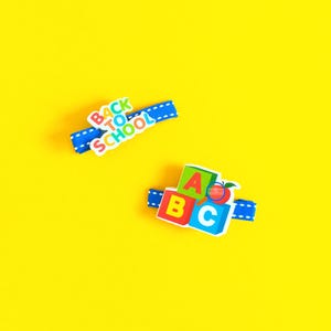 May include: Two blue hair clips with school-themed designs on a yellow background. One clip says "Back to School" in white and colourful letters. The other clip has ABC blocks and a red apple.